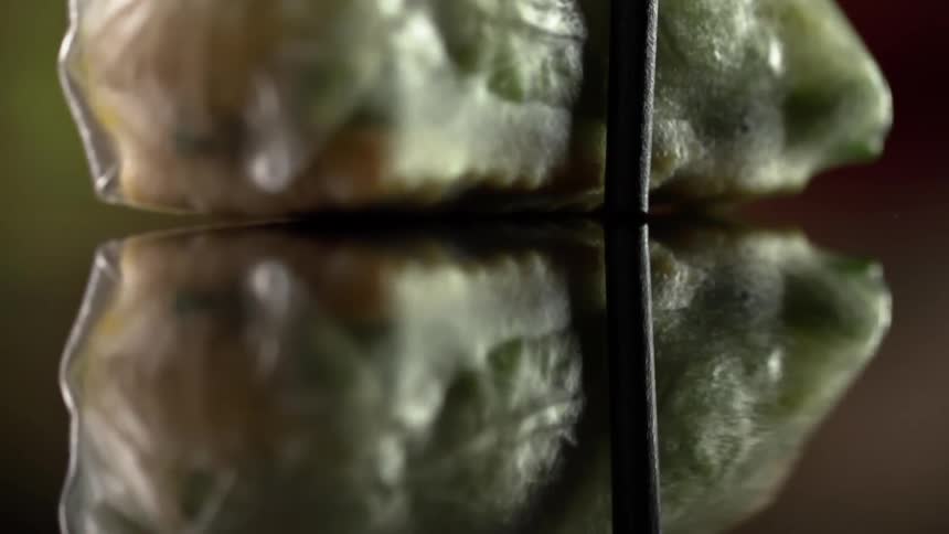 A tightly wrapped spring roll is placed on a highly reflective surface using chopsticks