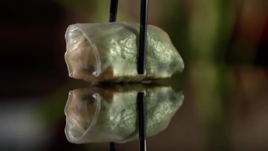 Black chopsticks lift a spring roll off a mirror like surface