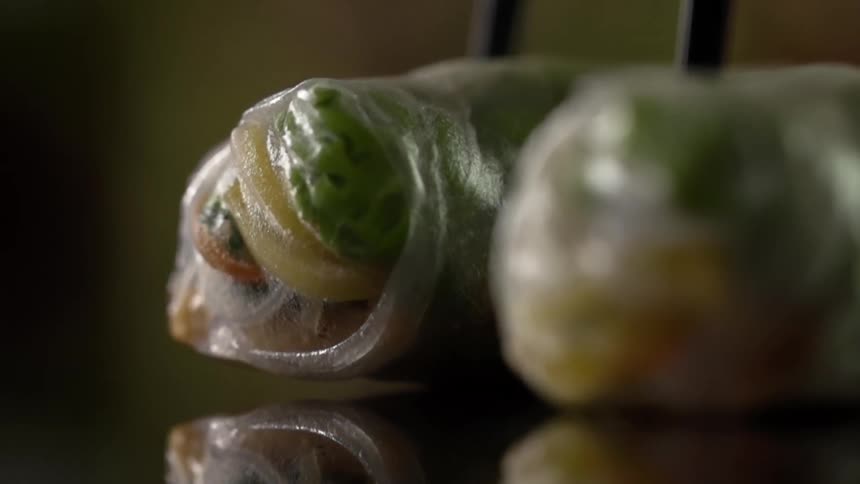 A spring roll is placed next to another one onto a highly reflective surface using a pair of chopsticks