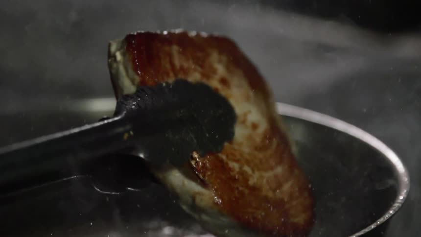 A charred salmon steak is held with a pair of tongs over a pot of boiling oil