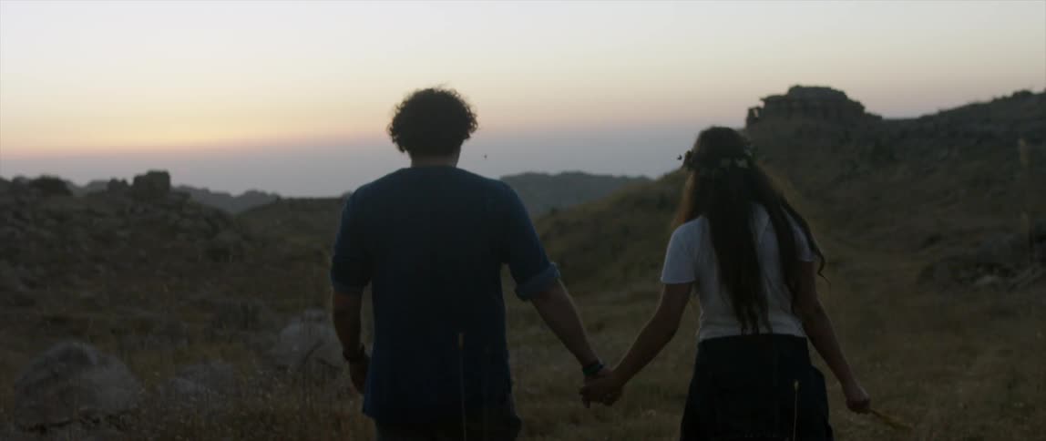 Two people holding hands walk over some hills at sunset