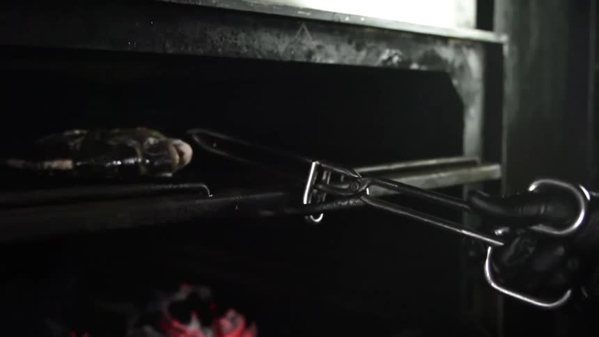 A gloved hand uses tongs to push a fish on a hot grate into an indoor grill with glowing coals