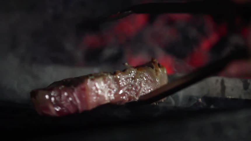 A steak is placed on a smoking grill with tongs