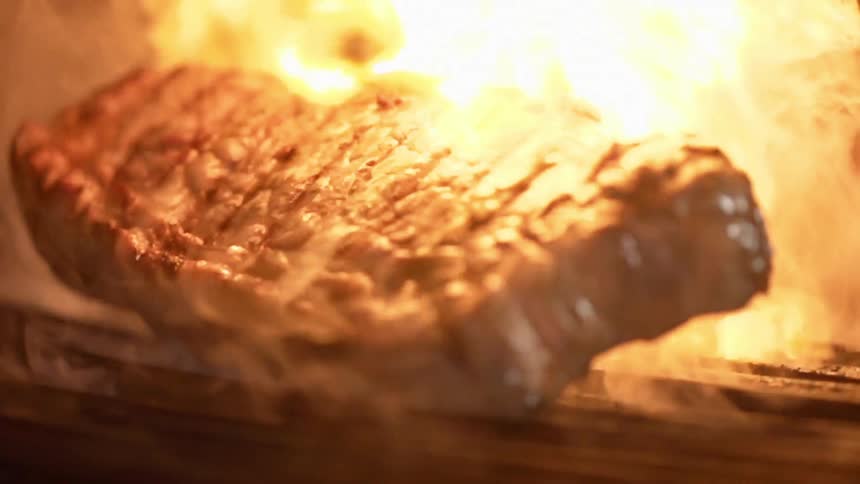 A cooked piece of steak drops down onto a flaming grill