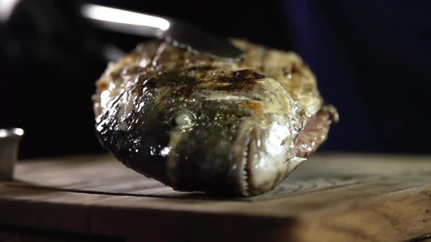 A cooked whole fish is placed down on a wooden cutting board with tongs