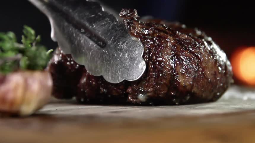 A thick and moist piece of grilled steak is gently placed on a cutting board using metal tongs