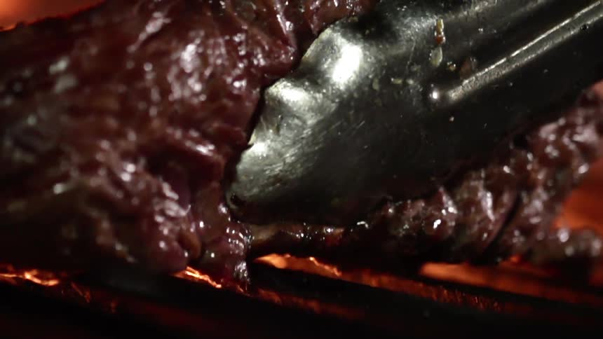 Metal tongs lift a piece of cooked steak off of a flame grill