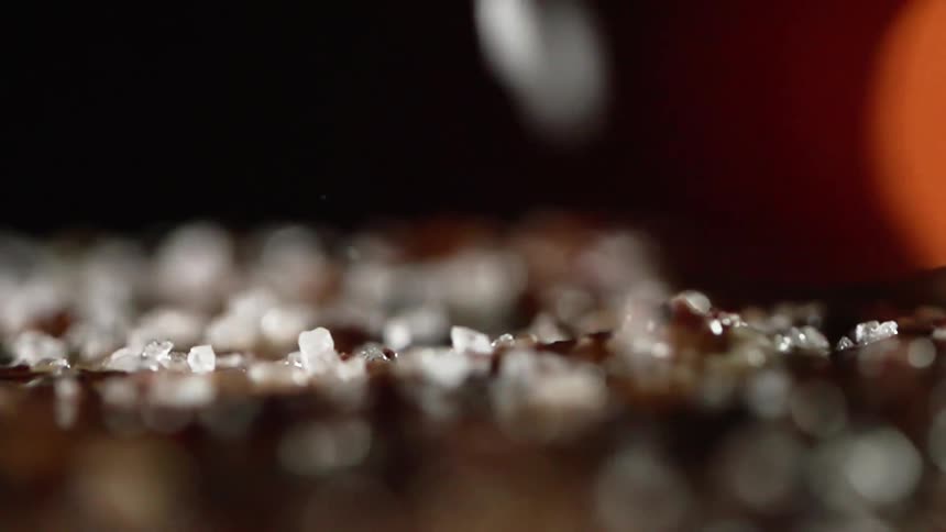 Coarsely ground salt crystals are sprinkled onto a food preparation surface as a flame flickers in the background