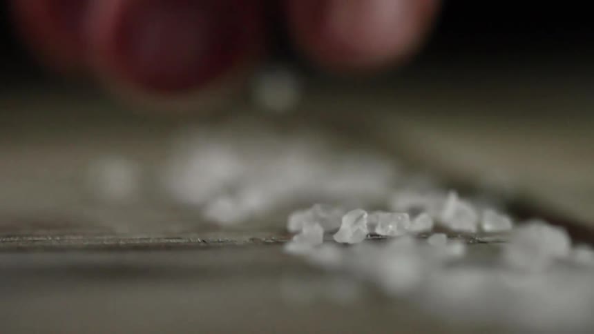 A person slowly sprinkle coarsely ground salt crystals onto a cutting board in a straight line