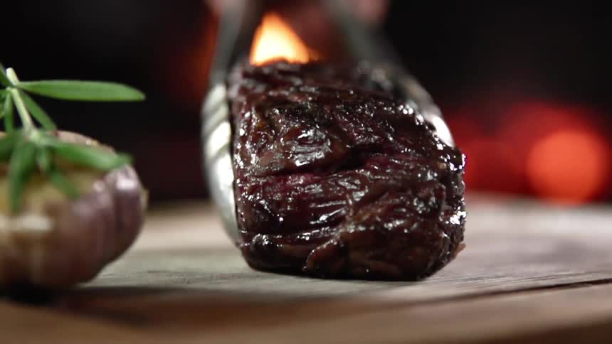 Person uses metal tongs to place cooked beef on wooden surface near rosemary topped vegetable