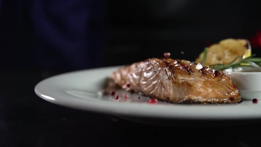 Pomegranate arils fall on grilled salmon on a white plate standing out against the dark background