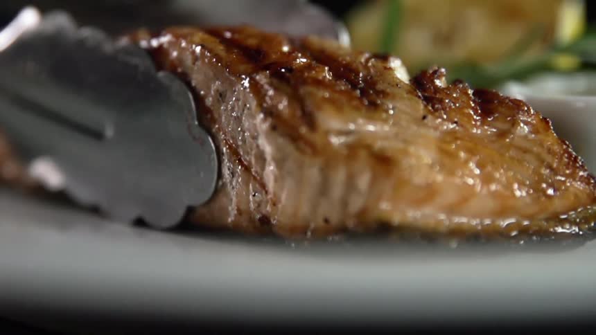 Close up of grilled salmon is being put on a plate by a pair of tongs