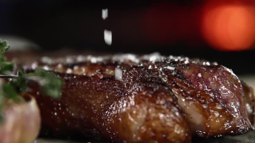 Salt and seasoning is sprinkled over the top of a grilled beef steak