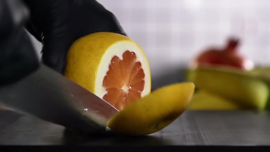 The cook wearing black gloves cuts a grapefruit with a knife