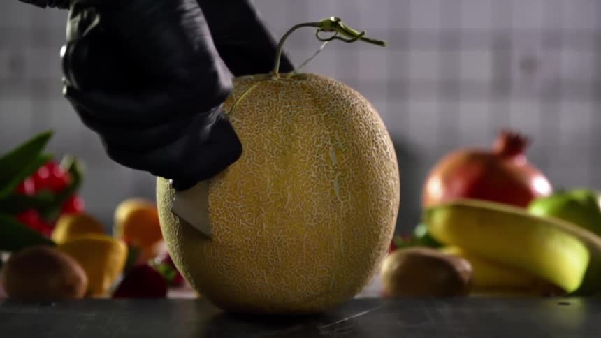 The cook wearing black gloves cuts a melon in half