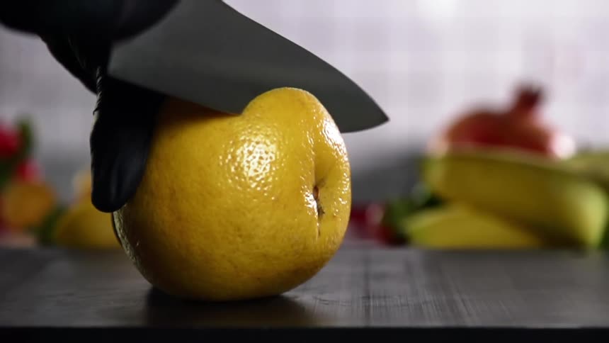 A person wearing a black disposable glove holds an orange and slices it with a sharp knife