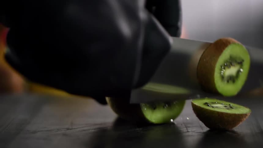 A kiwi being sliced with knife by a person wearing black gloves