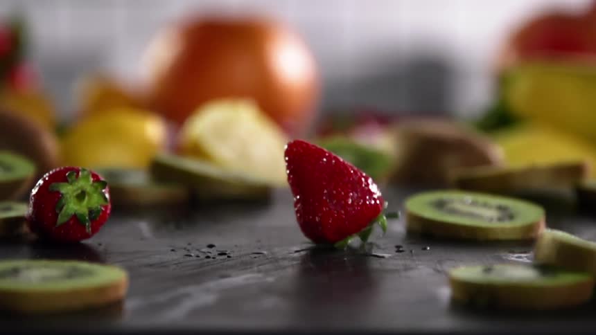 Three freshly washed strawberries are dropped onto a cutting board lined with many other pieces of sliced fruit