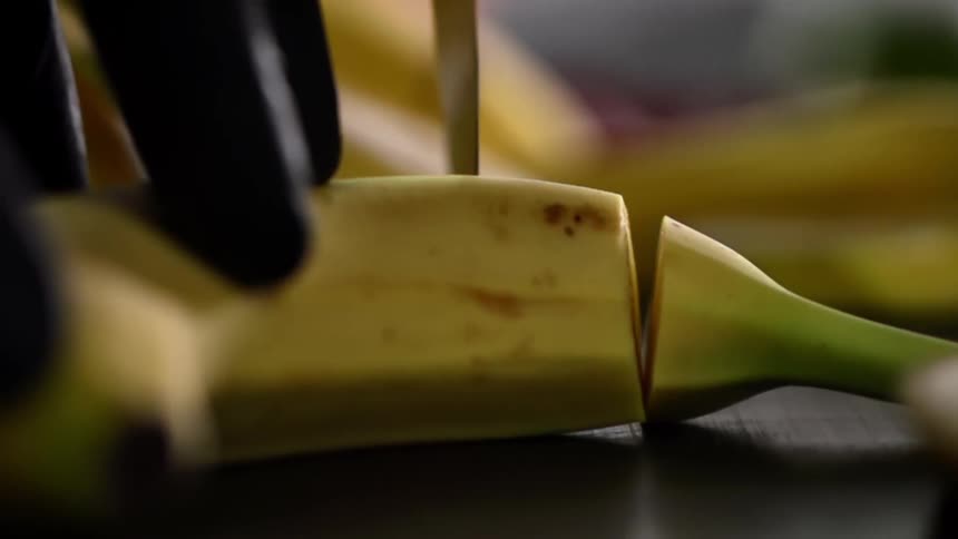 A person holds an unpeeled banana and carefully cutting slices of it using a sharp knife