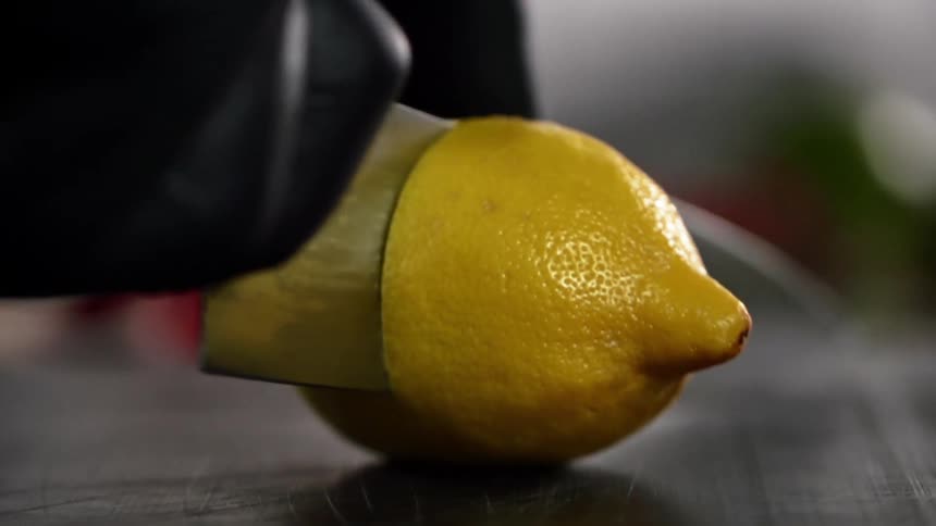 Cook wearing black gloves cuts a lemon in half with a knife on a cutting board