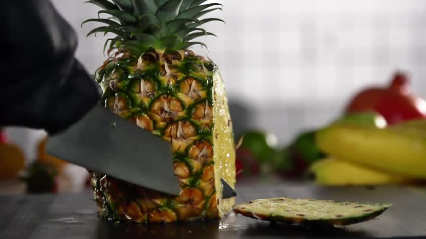 A person wearing a black gloves slices chunks off of a pineapple