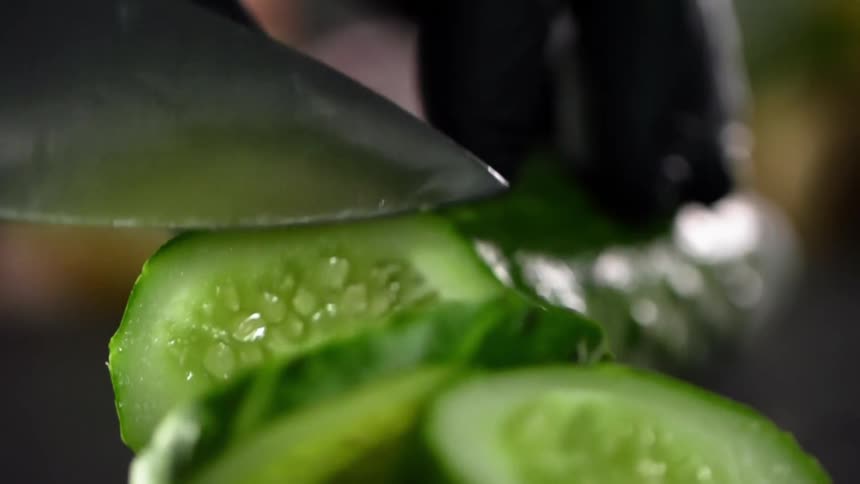 Close up of hands wearing black gloves cutting a long thin dark green cucumber with a sharp knife