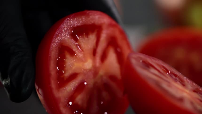 The cook wearing black gloves cuts a tomato in half