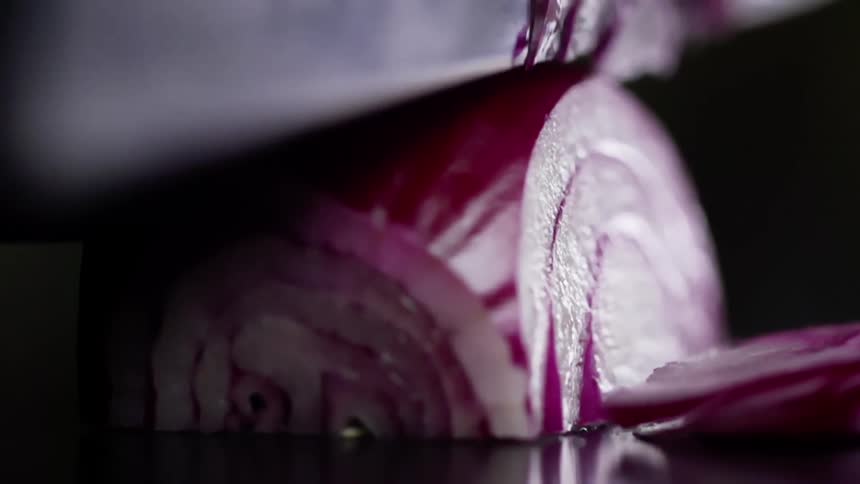 A large kitchen knife is used to cut very thin slices of a red onion