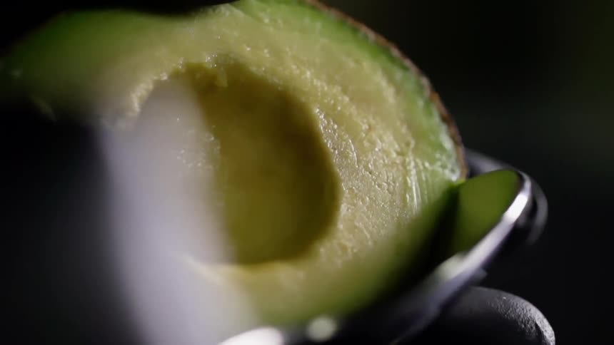 A person uses a spoon to scoop the ripe avocado from its skin