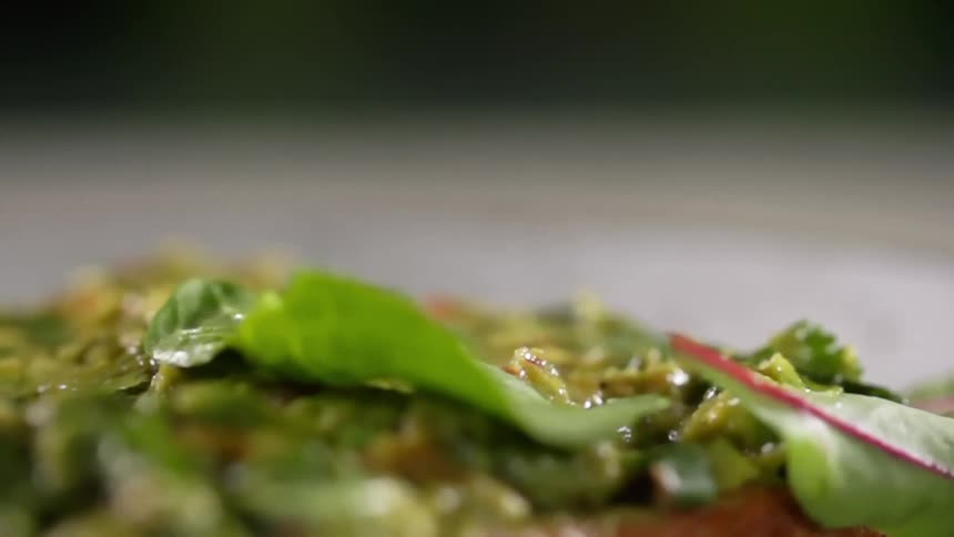 Green leafs fall over a slice of bread covered in guacamole in a white ceramic dish