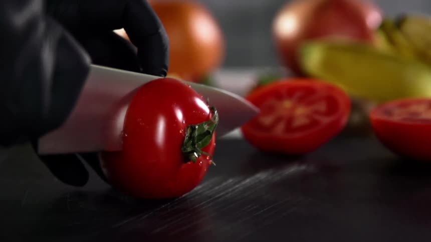 A small washed tomato is carefully sliced by a person wearing black disposable gloves