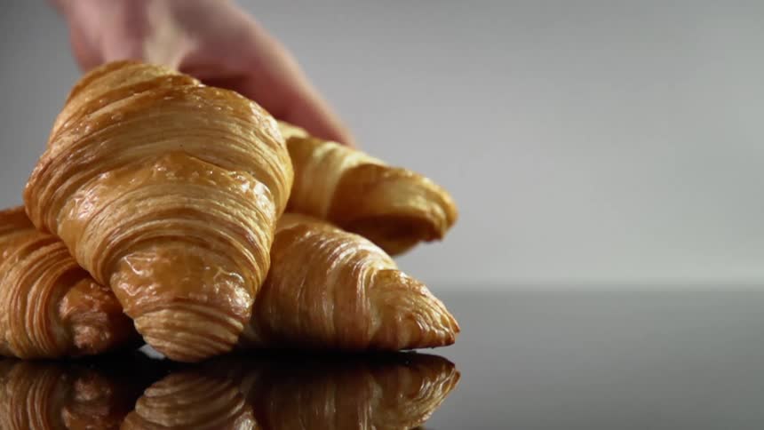 A person slowly lifts a croissant from a small pile of them stacked on a highly reflective counter top