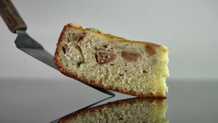 A slice of coffee cake is gently placed on a reflective glass surface by a metal spatula