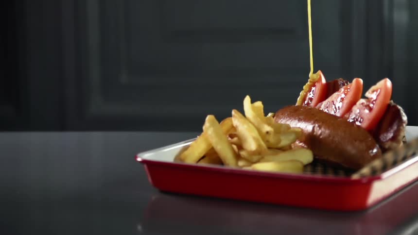 Mustard pouring over a tomato and sausage sandwich next to french fries