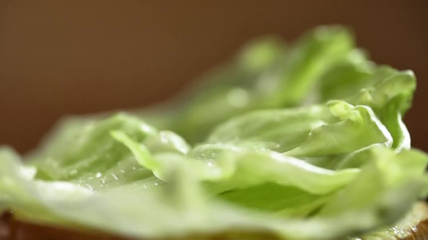 A stack of fresh green lettuce falls onto a sauce covered burger bun