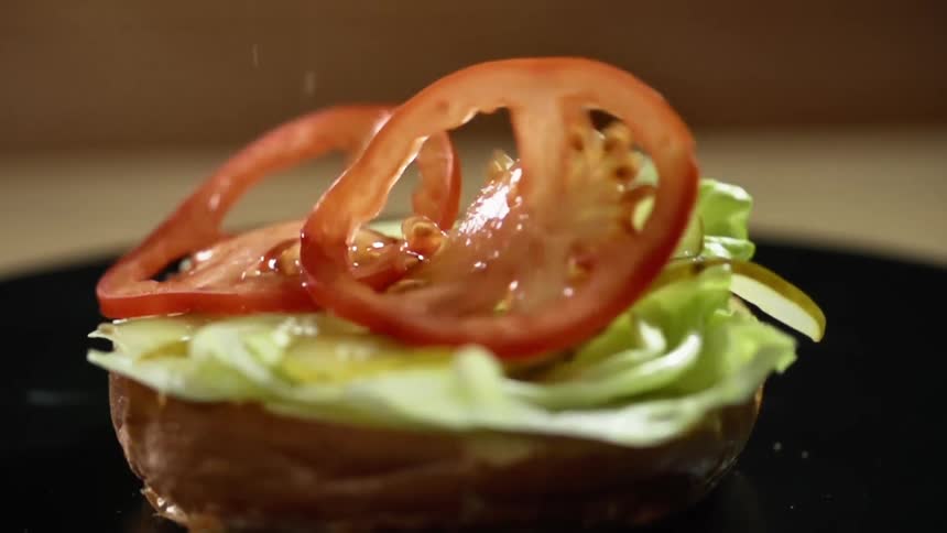 Red tomato slices fall over lettuce and pickles in the bottom half of a hamburger bun