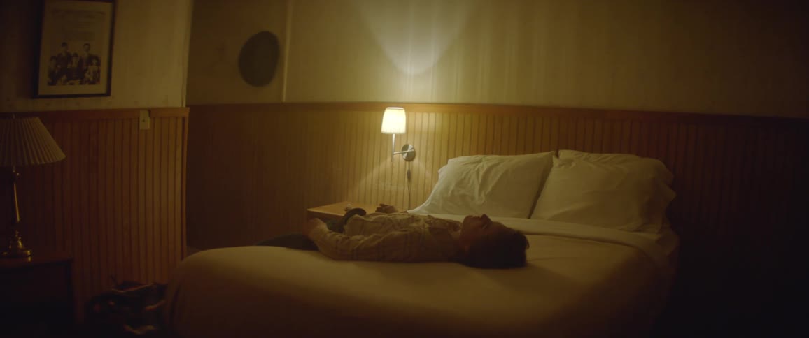 A man sitting on a motel bed leans back and lies down