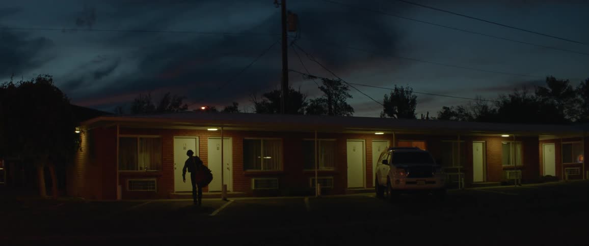 A man is carrying a saddle and gear towards a motel room at dusk