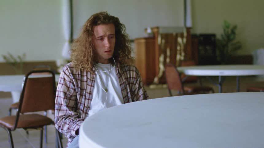 A man sits in a banquet chair by himself listening to earbuds looking sad and depressed