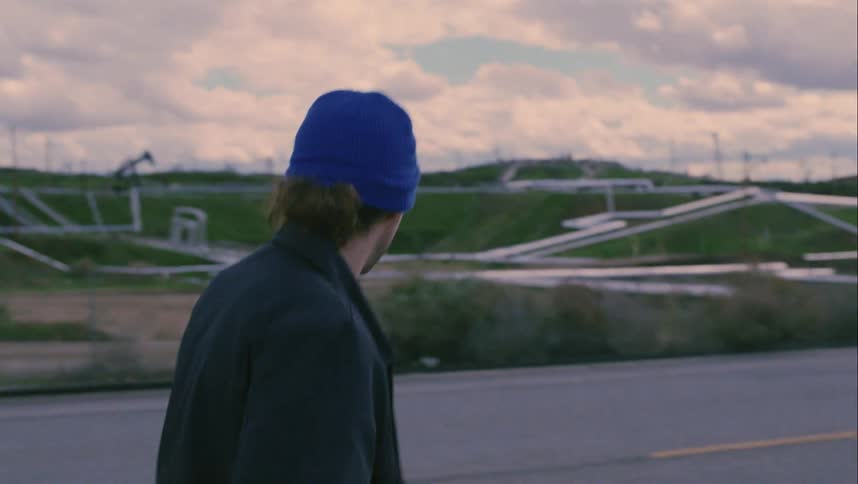 Man walks along road past field containing oil pump jack and pipes scanning area nervously