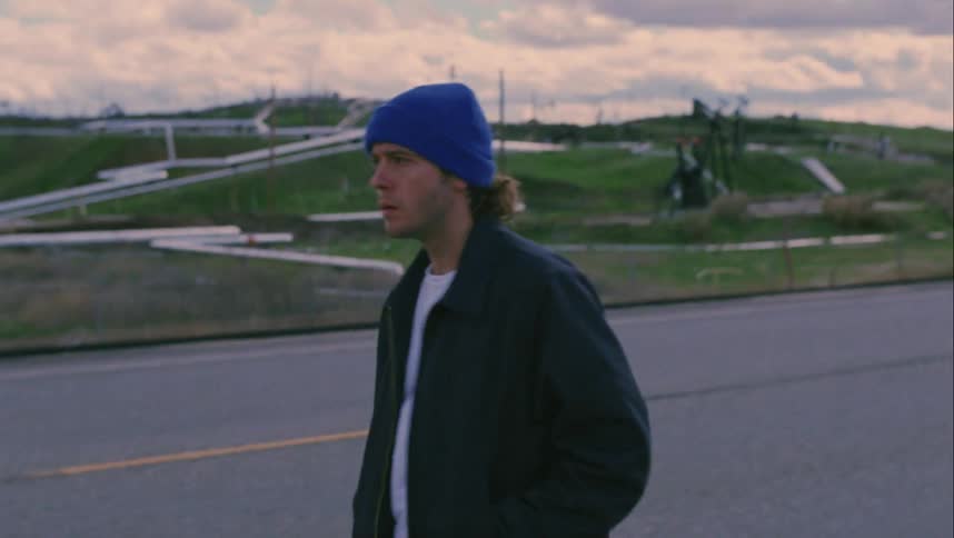 A man with a hat walking in the side of a road with the sky and clouds in the background