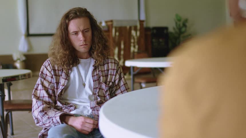 A young man slouches in a chair and frowns slightly as he listens to the woman talking to him