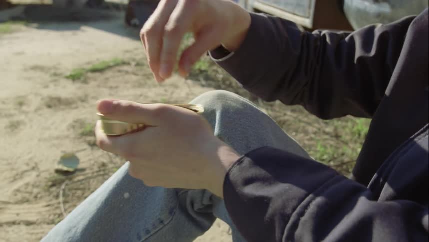 Man sitting on ground near car opens tin of sardines and eats