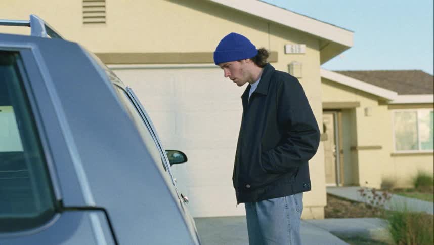 A teenager standing outside a house looks in a car and then backs away