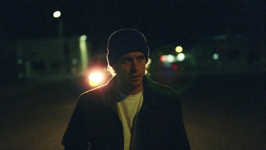 Man wearing knit hat looks around as vehicle with lit headlights drives past at night