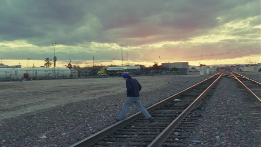 A man in a blue jacket and cap walks across a pair of parallel train tracks in a small town