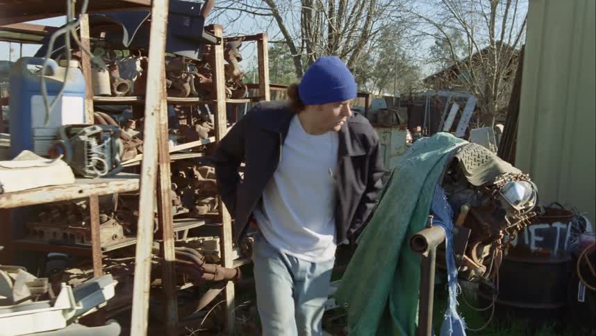 A man in a hat walking through a junkyard with trees and the sky in the background