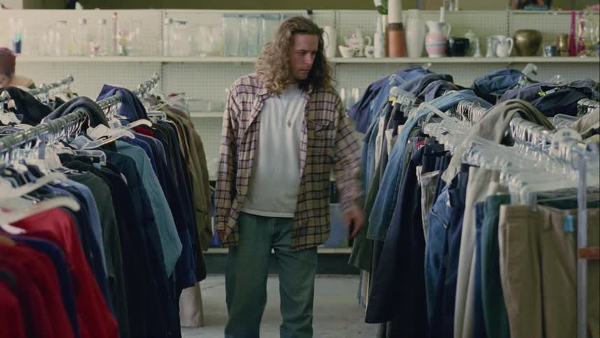 A young man with long curly hair looks through racks of hanging clothes inside of a retail store