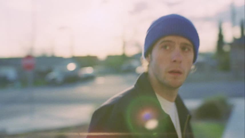 A man with a hat standing then walking in a neighborhood with houses trees and vehicles