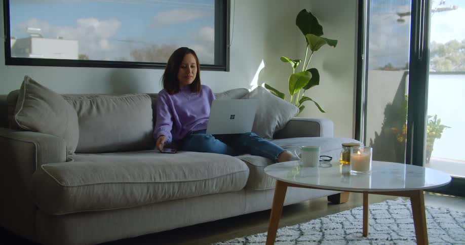 Woman sitting in a couch in the living room talks to the phone while typing on a laptop